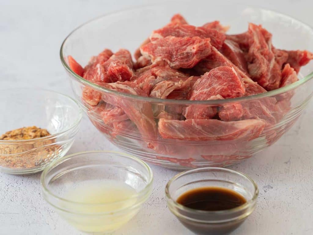 Slices of flank steak in a glass bowl. Seasonings, oil and worchestire sauce in mini glass bowls.