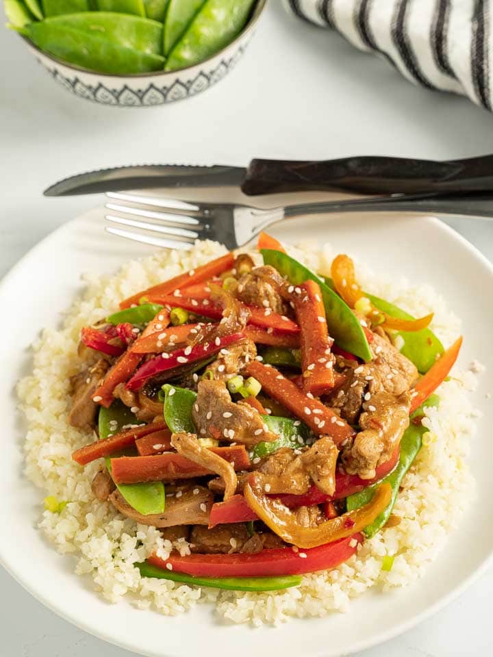 keto chicken stir fry with cauliflower rice on white plate with knife, fork and striped napkin

