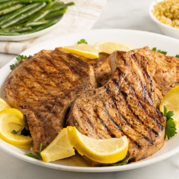 lemon pepper pork chops on white platter with lemon, green beans.