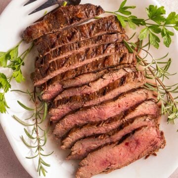 sliced flank steak on white platter with herbs