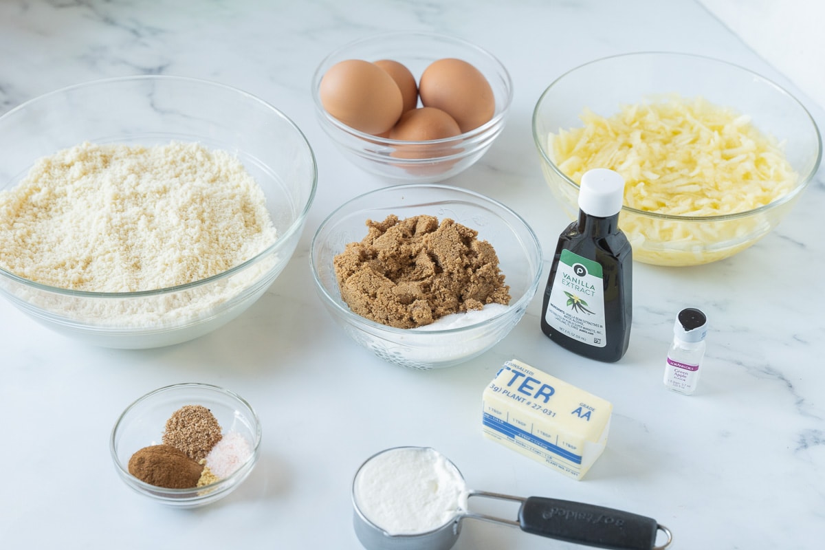 Grated apples, brown sugar, eggs, almond flour, spices in glass bowls, vanilla, butter, sour cream.