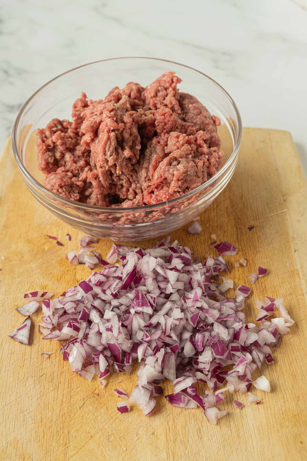 ground beef in glass bowl, chopped red onion on cutting board.