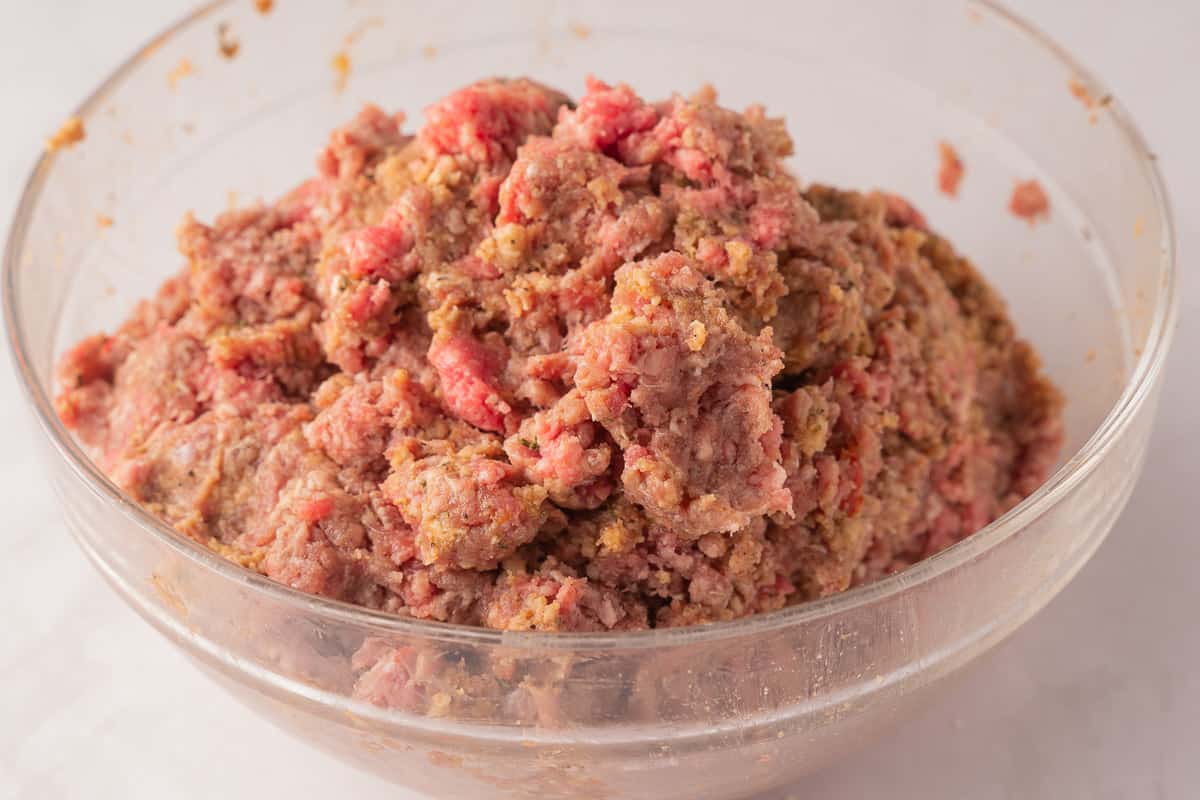 Ground beef combined with panko, seasonings, ketchup, chili sauce in glass bowl.