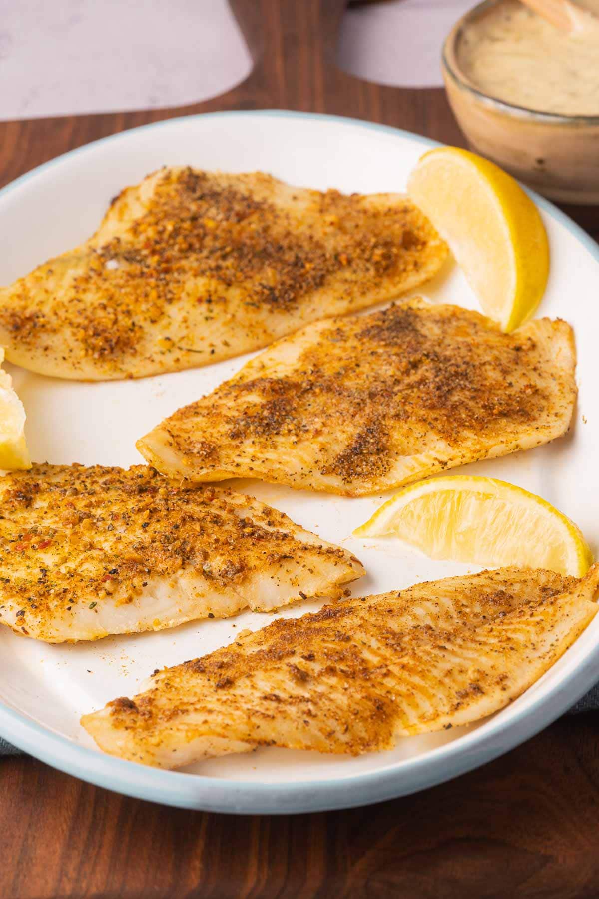 Broiled flounder on white platter with lemon slices and cup of tartar sauce.