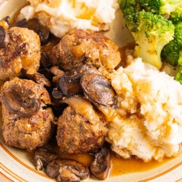 A beige plate with salisbury steak meatballs, mashed potatoes, gravy and broccoli.