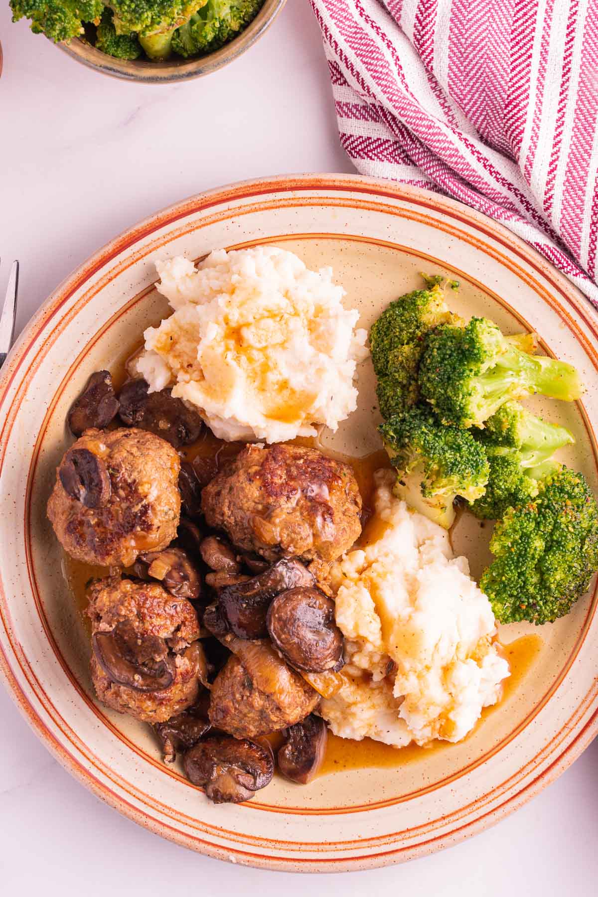 Slow Cooker Salisbury Steak Meatballs, gravy, mashed potatoes, broccoli on plate with striped napkin.