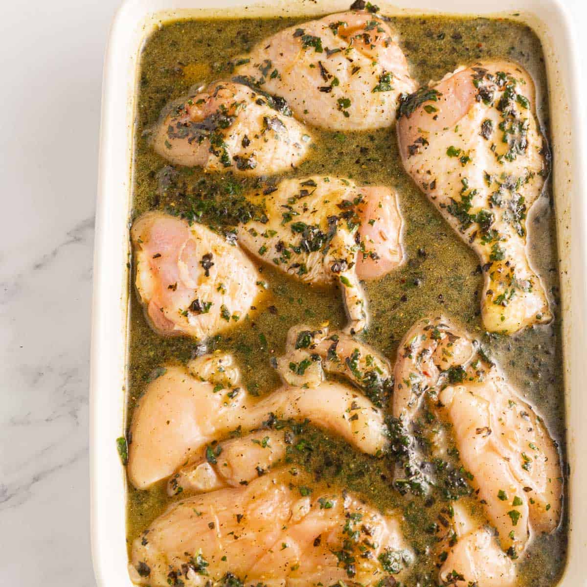 Chicken legs and breasts in basil chicken marinade in a white casserole.
