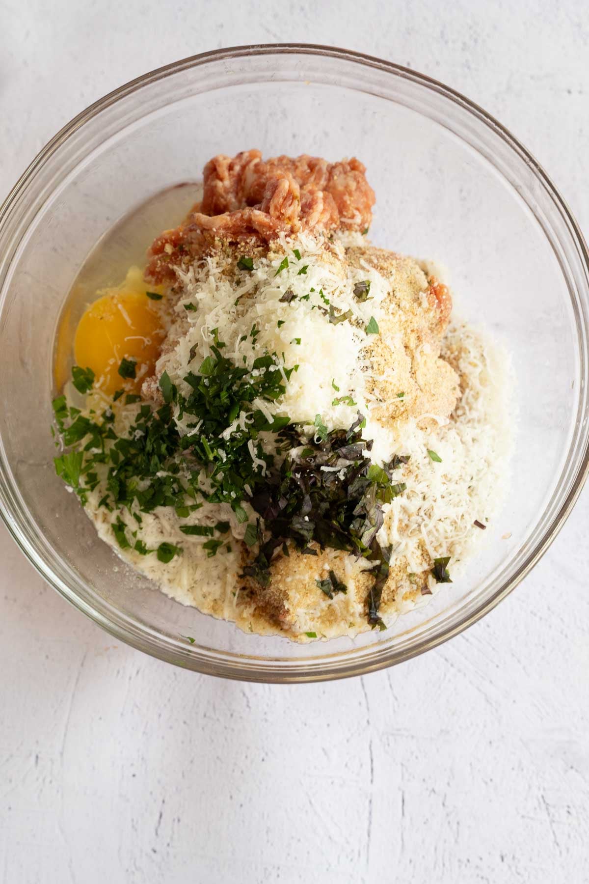 Breadcrumbs, ground sausage, seasonings, egg in glass bowl.
