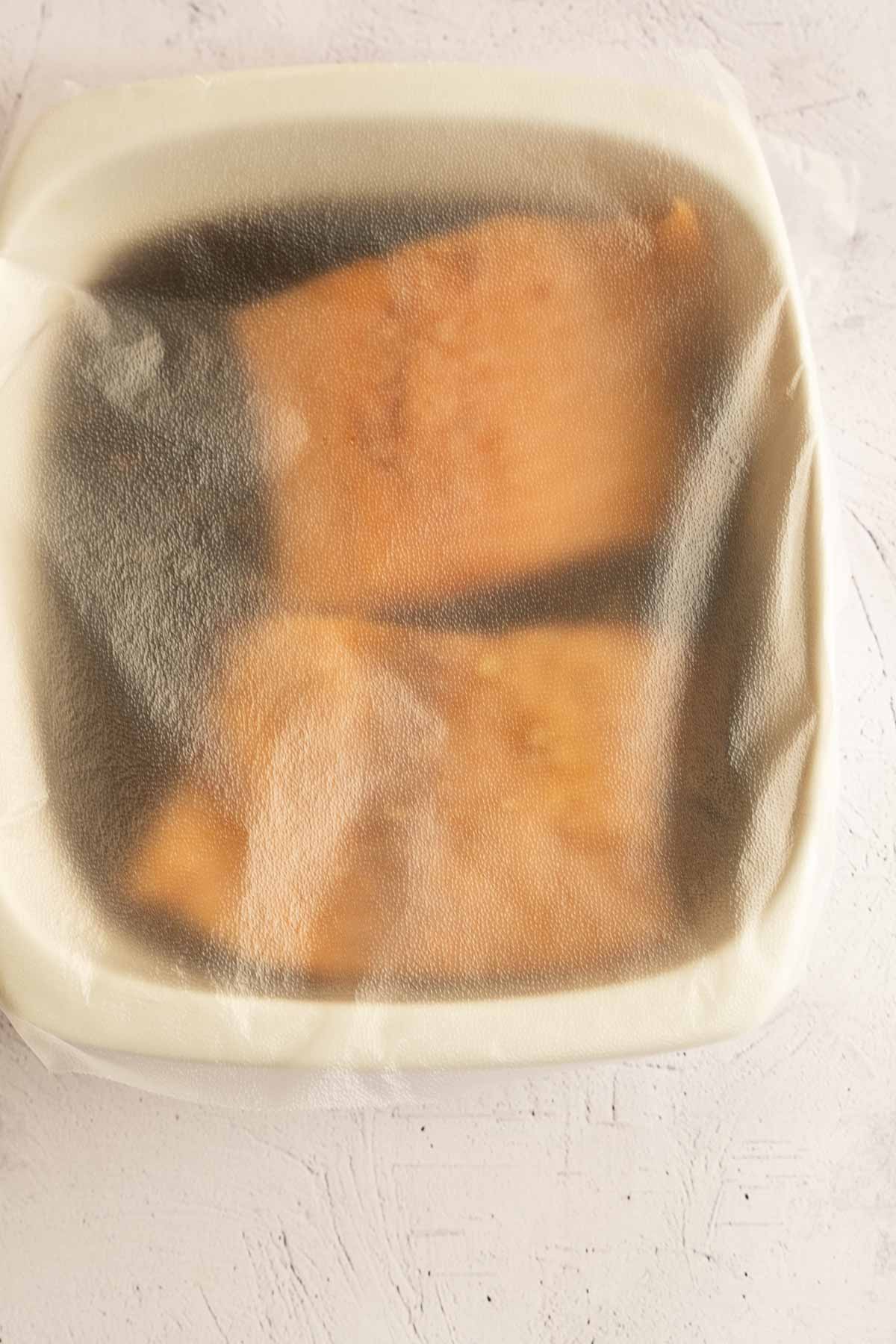 Marinated salmon fillets in bowl, covered with plastic wrap.